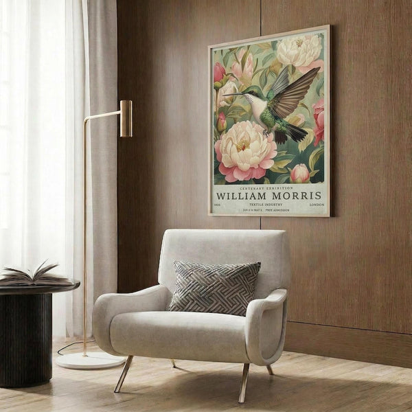 May include: Framed botanical poster featuring a green hummingbird with spread wings among pink and white peonies, displayed above a beige upholstered armchair with geometric pillow in a modern living room with wood paneling, brass floor lamp, and side table with open book.