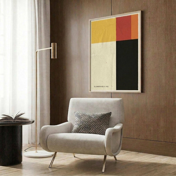 May include: Modern living room with beige armchair and geometric pillow, brass floor lamp, dark round side table with open book, and wood panel wall displaying framed abstract art print with yellow, red, orange, cream, and black color blocks in minimalist composition.