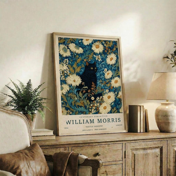 May include: Framed poster displaying blue floral pattern with white daisies and black cat silhouette, featuring William Morris exhibition text at bottom. Light wood frame sits on rustic wooden sideboard next to fern plant, ceramic vases, and books. Cream armchair with brown leather pillow visible in corner of bright, naturally-lit room with white walls.