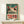 May include: A vintage-style poster featuring a white frog with large red-rimmed eyes and red spots against a teal background, with "TOKYO" text and Japanese characters. The artwork has a weathered, retro appearance in a thin wooden frame, displayed on a white shelf with a small wrapped gift box beside it.