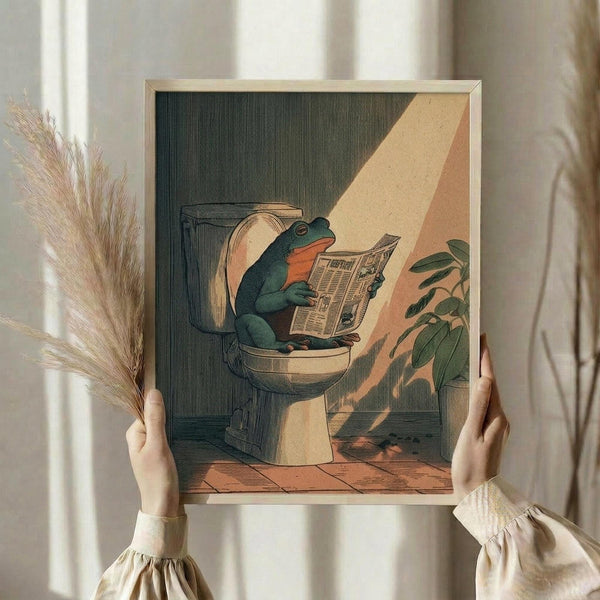 May include: Hands holding a framed vintage-style illustration of a green frog with orange throat sitting on a toilet reading a newspaper in a tiled bathroom with sunlight streaming through. The artwork has an antique engraving aesthetic with muted green, orange and cream tones. Light wooden frame against neutral curtains with dried pampas grass visible on left side.