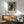 May include: Framed poster featuring a black cat surrounded by blue floral wallpaper with white daisies and green foliage, displayed above a wooden sideboard with table lamp in a mid-century modern living room setting with sage green upholstered chair and round coffee table.