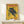 May include: Vintage-style poster with yellow background featuring a black panther perched on tree branches, red "TOKYO" text at top and "JAPAN WEEK" at bottom, Japanese characters on left side, displayed in thin wooden frame on light wood surface next to small white wrapped gift with natural twine and sprig.
