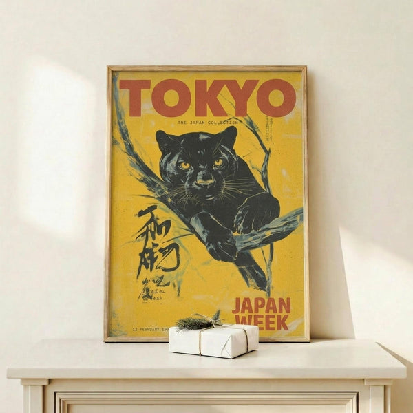 May include: Vintage-style poster with yellow background featuring a black panther perched on tree branches, red "TOKYO" text at top and "JAPAN WEEK" at bottom, Japanese characters on left side, displayed in thin wooden frame on light wood surface next to small white wrapped gift with natural twine and sprig.