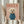 May include: Framed poster with pink background showing a small white fluffy dog wearing a blue turtleneck sweater, with "DOGUE" text in orange letters above. Person in cream ruffled sleeves holds the light wood frame alongside dried wheat stems against white curtained window.