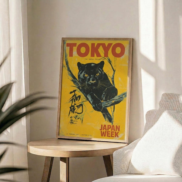 May include: Framed vintage-style poster with yellow background featuring black panther on tree branch, red "TOKYO" text at top and "JAPAN WEEK" at bottom, Japanese characters on left side, displayed on round wooden side table next to beige upholstered chair with white throw pillow, plant leaves visible on left, natural sunlight casting shadows on white wall