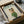 May include: Framed poster print in black frame being unpacked from cardboard shipping box with protective wrapping. Features white poodle wearing green knit sweater, sunglasses, and colorful plaid head scarf with "DOGUE" text in green letters above. Cream colored background with fashion magazine aesthetic. Surrounded by brown packing paper and tissue paper on neutral surface.