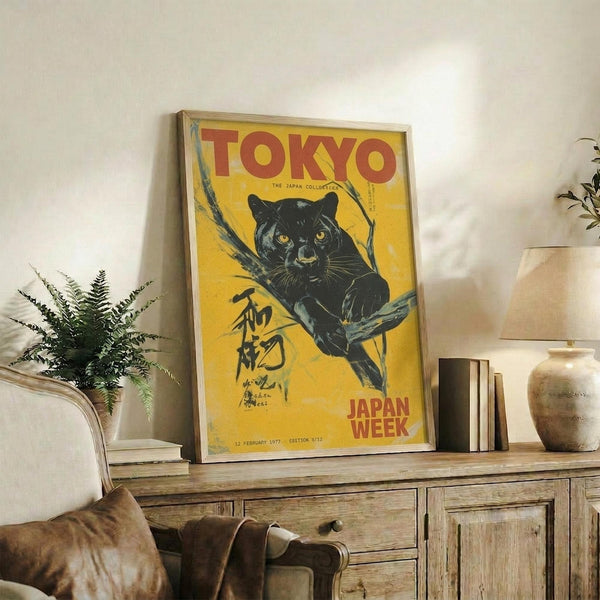 May include: Framed vintage-style yellow poster featuring a black panther on tree branches with Japanese text and "TOKYO" and "JAPAN WEEK" in red lettering, displayed on rustic wooden sideboard alongside fern plant, ceramic vase, stacked books, and beige armchair in neutral-toned living room setting