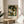 May include: Framed botanical poster featuring two white swans with orange beaks surrounded by cream peonies and green foliage on dark background, displayed on round wooden side table next to beige upholstered chair in sunlit room with white curtains and plant shadows