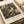 May include: Vintage-style poster print featuring a colorful bird with cream and rust-colored plumage perched among ornate botanical foliage with orange and yellow flowers on dark blue background. Print shows exhibition text below the main illustration and appears rolled in kraft paper packaging on white surface with natural lighting.