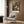 May include: vintage-style botanical poster featuring colorful bird among flowers and foliage, displayed on wood-paneled wall above cream upholstered chair with geometric pillow, brass floor lamp and side table with open book nearby in modern interior setting