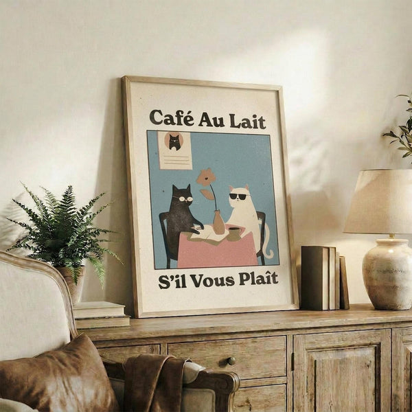 May include: Framed vintage-style poster showing two cats sitting at a cafe table with coffee cups, featuring French text "Café Au Lait" and "S'il Vous Plait" in black lettering. The illustration uses muted blue, pink, and cream colors with a minimalist art style. Display shows poster leaning against white wall on rustic wooden sideboard alongside fern plant, books, ceramic vase, and brown leather chair visible in foreground of cozy living room setting.