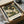 May include: Framed vintage-style poster featuring two white swans with orange beaks surrounded by lush green foliage and cream-colored flowers, displayed in a black frame inside an open cardboard shipping box with protective tissue paper wrapping on a white surface.