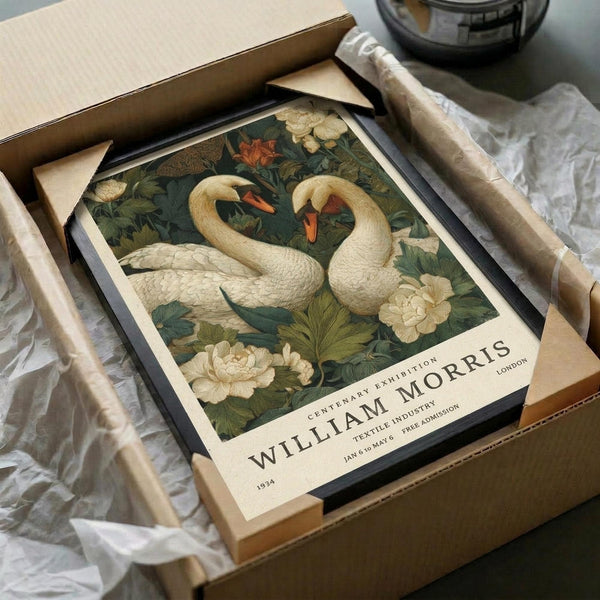 May include: Framed vintage-style poster featuring two white swans with orange beaks surrounded by lush green foliage and cream-colored flowers, displayed in a black frame inside an open cardboard shipping box with protective tissue paper wrapping on a white surface.