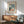 May include: Framed poster print featuring two cats at a small table with coffee cups, displayed above a mid-century modern wooden credenza with white ceramic table lamp. Green upholstered armchair with wooden frame sits beside a round wooden side table. Warm sunlight creates geometric shadows on white wall. Print shows vintage-style illustration with French text in cream and teal color palette with wooden frame.
