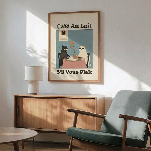 May include: Framed poster print featuring two cats at a small table with coffee cups, displayed above a mid-century modern wooden credenza with white ceramic table lamp. Green upholstered armchair with wooden frame sits beside a round wooden side table. Warm sunlight creates geometric shadows on white wall. Print shows vintage-style illustration with French text in cream and teal color palette with wooden frame.