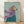 May include: Framed painting of a tan Italian Greyhound wearing a purple headscarf and teal coat against a sage green background, displayed in a light wood frame leaning against white walls on wooden flooring with soft natural lighting creating diagonal shadows