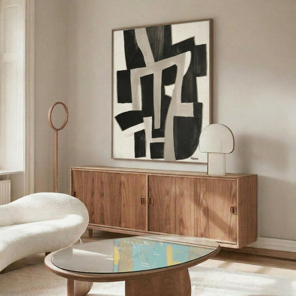May include: Abstract black and white geometric poster with bold angular shapes displayed above a mid-century modern walnut credenza. Room features cream walls, white upholstered chair, oval glass coffee table, and contemporary lighting. Artwork shows irregular black forms on light background in natural wood frame.