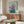 May include: Framed artwork showing an Italian Greyhound wearing a purple headscarf and teal clothing against a mint green background, displayed above a mid-century modern walnut credenza with white table lamp and brass floor lamp, white curved chair and glass coffee table visible in minimalist living room setting.