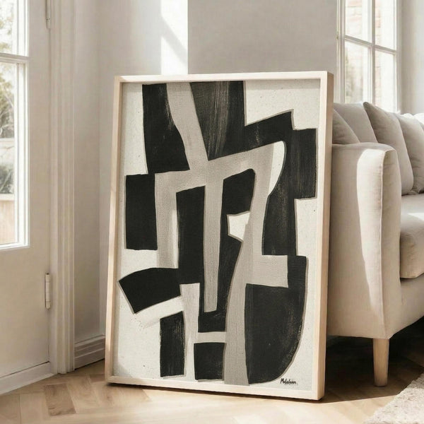 May include: Large framed abstract artwork leaning against white wall next to beige sofa. Black and white geometric composition with bold angular shapes and intersecting lines creates dynamic visual rhythm. Natural wood frame complements neutral living room setting with hardwood floors and bright windows.