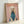May include: Framed poster print leaning against white wall corner showing poodle with fluffy cream-colored coat wearing teal turtleneck sweater on soft pink background with "DOGUE" text in orange serif letters at top, displayed in light wood frame