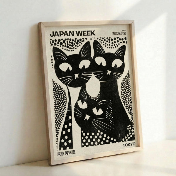 May include: Framed vintage-style poster featuring three stylized black cats with white facial markings and whiskers, arranged in a family group against cream background with polka dot patterns. Text reads "JAPAN WEEK" at top with "1984" and Japanese characters, "TOKYO" at bottom. Poster displayed in light wood frame leaning against white wall.