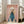 May include: Framed poster featuring a white poodle with fluffy curly fur wearing a teal blue turtleneck sweater against a soft pink background, with "DOGUE" text in orange letters at the top. The artwork is displayed in a light wood frame, leaning against a white wall near beige upholstered furniture and tall windows with natural lighting in a modern living room setting.