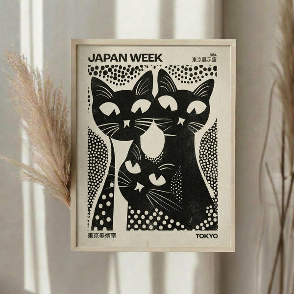 May include: A framed vintage-style black and white poster featuring three stylized cats in bold graphic design, displayed on a white wall next to dried pampas grass in neutral tones. The artwork shows geometric patterns and Japanese text, housed in a light wooden frame against soft natural lighting and cream curtains.
