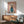 May include: Framed poster print on white wall showing white poodle wearing teal blue turtleneck sweater against pink gradient background with "DOGUE" text at top, displayed above wooden sideboard with white table lamp and mid-century modern sage green upholstered armchair in sunlit room