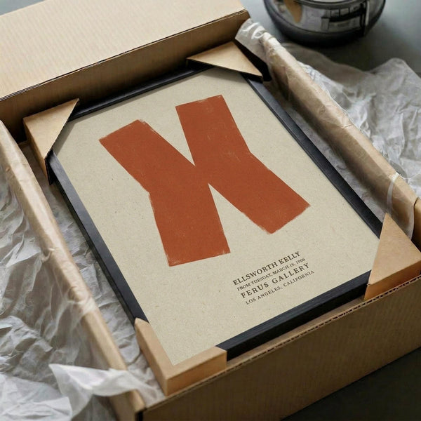 May include: Framed minimalist poster featuring a large orange letter "N" on cream background with small gallery text below, displayed in black frame inside cardboard shipping box with tissue paper wrapping and wooden frame corners visible