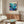 May include: Framed poster with vibrant underwater coral reef scene in blues, purples, pinks and oranges, featuring Japanese text and "JAPAN 1984" with museum attribution, displayed above mid-century walnut credenza in modern living room with white curved chair, brass floor lamp, and glass-top coffee table on light wooden floors