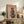 May include: Framed poster featuring a whippet dog wearing a gray knit beanie and fuzzy brown sweater on pink background with "DOGUE" text in red letters, displayed on rustic wooden console table alongside books, ceramic vases, fern plant, and cream upholstered chair in cozy living room setting with white walls.