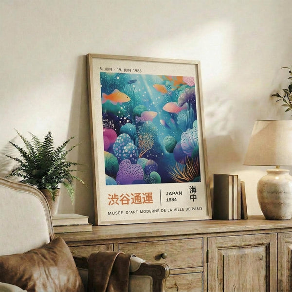 May include: Vintage-style museum poster with underwater coral reef scene in vibrant blues, purples, and oranges, featuring fish and sea life. Text shows dates and Japanese characters. Displayed in light wood frame on rustic wooden sideboard with books, ceramic vases, fern plant, and cream upholstered chair with brown throw pillow in cozy living room setting.