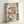 May include: Framed art print leaning against white wall showing three red spotted fish with large black eyes swimming among green botanical elements, white flowers, and scattered dots on cream background. Natural wood frame, soft lighting creates gentle shadows on wall.