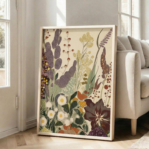 May include: Large framed botanical artwork leaning against white wall next to beige sofa in bright living room. The poster features stylized wildflowers and plants in dotted patterns with white daisies, purple lavender spikes, yellow blooms, and rust-colored foliage against cream background. Natural wood frame, hardwood floors visible.