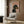 May include: Framed poster showing a black and tan dachshund wearing a pearl collar against a pink background with "DOGUE" text in orange letters, displayed on wood paneled wall above cream upholstered chair with geometric patterned pillow, brass floor lamp and side table with open book nearby