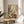 May include: Framed botanical art print displayed on round wooden side table next to beige upholstered chair. Artwork features stylized wildflowers and plants in dotted, textured style with white daisies, purple lupines, yellow blooms, and various grasses in earthy tones. Natural lighting creates soft shadows across neutral room setting with plant leaves visible in corner.