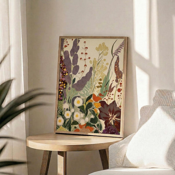 May include: Framed botanical art print displayed on round wooden side table next to beige upholstered chair. Artwork features stylized wildflowers and plants in dotted, textured style with white daisies, purple lupines, yellow blooms, and various grasses in earthy tones. Natural lighting creates soft shadows across neutral room setting with plant leaves visible in corner.