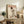 May include: Framed poster featuring a white Maltese dog wearing a black top hat with red band and cream knit sweater, styled like a vintage magazine cover with "DOGUE" in red letters and Christmas ornaments at the bottom. The light wood frame sits on a rustic wooden credenza beside a fern plant, books, and ceramic vase in a cozy living room setting with beige chair visible.