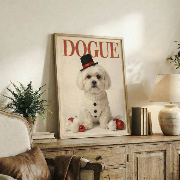 May include: Framed poster featuring a white Maltese dog wearing a black top hat with red band and cream knit sweater, styled like a vintage magazine cover with "DOGUE" in red letters and Christmas ornaments at the bottom. The light wood frame sits on a rustic wooden credenza beside a fern plant, books, and ceramic vase in a cozy living room setting with beige chair visible.