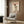 May include: Framed poster print hanging on wood-paneled wall above modern beige armchair with geometric pillow. Print shows white Maltese dog wearing black top hat with red band and cream cable-knit sweater, sitting with red and white Christmas ornaments. Red "DOGUE" text at top. Gold floor lamp and side table with open book visible in contemporary living room setting.