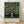 May include: Framed poster print leaning against white wall on light wood floor. Features vintage-style apple tree illustration with golden yellow apples, green foliage, and brown trunk against dark navy background. Exhibition-style poster with cream bottom section containing black text including "WILLIAM MORRIS" and exhibition details. Natural wood frame, soft shadows from window light visible on wall.