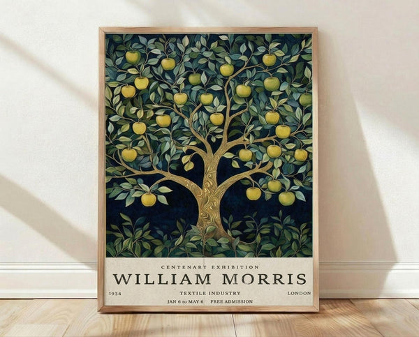 May include: Framed poster print leaning against white wall on light wood floor. Features vintage-style apple tree illustration with golden yellow apples, green foliage, and brown trunk against dark navy background. Exhibition-style poster with cream bottom section containing black text including "WILLIAM MORRIS" and exhibition details. Natural wood frame, soft shadows from window light visible on wall.