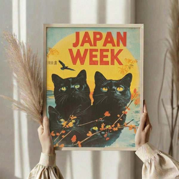 May include: Person in cream sweater holding framed vintage-style poster featuring three black cats with bright yellow eyes against sunset backdrop. Red text reads "JAPAN WEEK" above cats surrounded by orange cherry blossom branches. Flying bird silhouette and pagoda visible in background. Poster has warm yellow, orange, and teal color palette in thin gold frame. Dried pampas grass stems visible on left side.