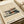 May include: Minimalist architectural poster featuring a modern two-story building with clean geometric lines, large windows, and earth-tone color palette of beige, orange, and dark green. The vintage-style print sits on cream paper with "BAUHAUS" and "INTERNATIONAL DESIGN MUSEUM" text at bottom, displayed on a neutral surface with rolled paper tubes nearby.