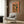 May include: Orange vintage-style travel poster featuring a woman surrounded by citrus fruits and leaves, displayed in light wood frame on wood paneled wall above cream upholstered armchair with geometric patterned pillow, brass floor lamp and dark side table with open book nearby