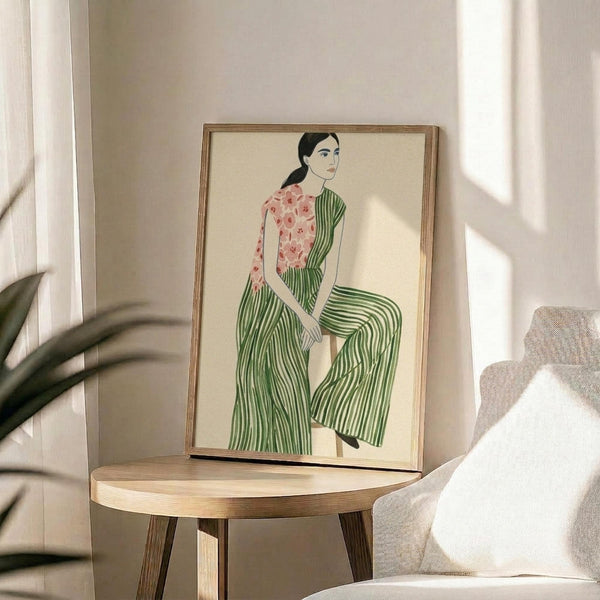 May include: Framed artwork displayed on round wooden side table next to beige upholstered chair with white pillow. Print shows woman with dark hair in ponytail wearing pink floral top and green striped wide-leg pants, sitting against cream background. Natural wood frame, soft sunlight streaming across neutral-toned room with plant leaves visible in corner.