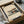 May include: Modern architectural print featuring a Bauhaus-style building with clean geometric lines in muted tones of beige, green, and orange, displayed in a black frame within a cardboard shipping box surrounded by protective tissue paper and wooden dowels for safe packaging.