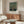May include: Framed William Morris exhibition poster featuring a decorative apple tree with yellow fruit and green foliage on dark background, displayed above a walnut wood credenza in modern living room with white curved chair, glass coffee table, and brass floor lamp