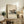 May include: Framed Bauhaus poster featuring modernist architectural illustration in muted beige, orange and green tones, displayed on rustic wooden console alongside fern plant, ceramic vases, and books in neutral living room setting with beige armchair and warm lighting